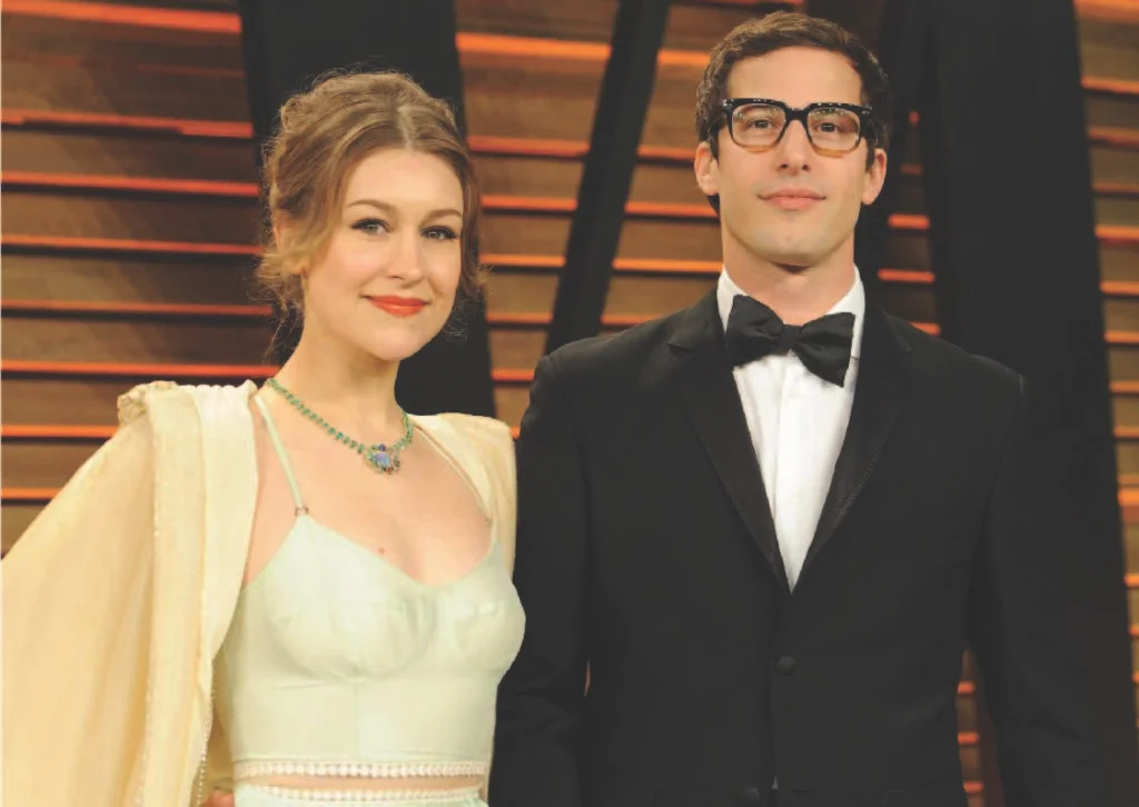 johanna samberg in a light green dress with a sheer cape and jewelry stands next to a man in a black tuxedo and glasses at an event.