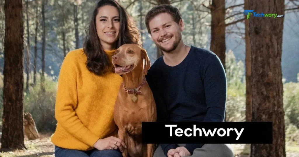Melanie Sergiev and a man smile with their dog in a forest setting.