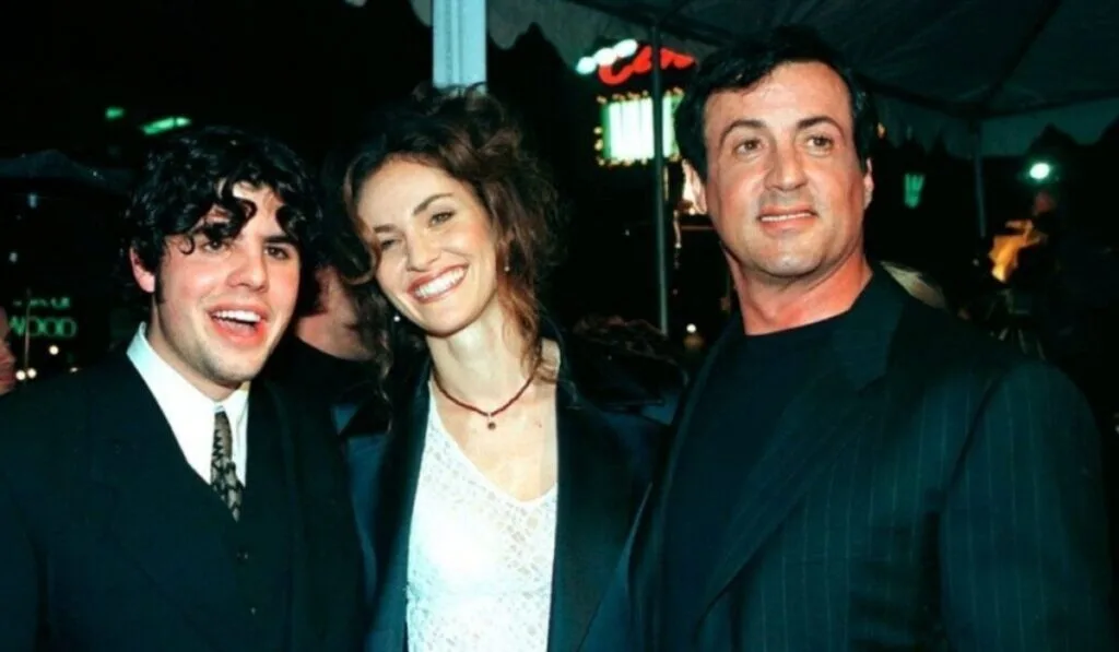 Three people pose for a photo outdoors at night. The man on the left is smiling widely, the woman in the center has a joyful expression, and the man on the right looks off-camera. They are dressed in formal attire, suggesting a celebratory or eventful setting.