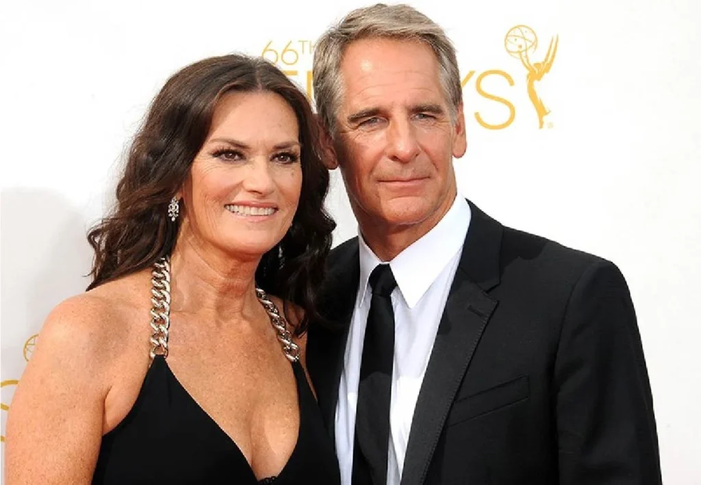 A smiling couple dressed elegantly on the red carpet. The woman wears a black dress with silver straps, the man wears a black suit. They appear relaxed and happy.